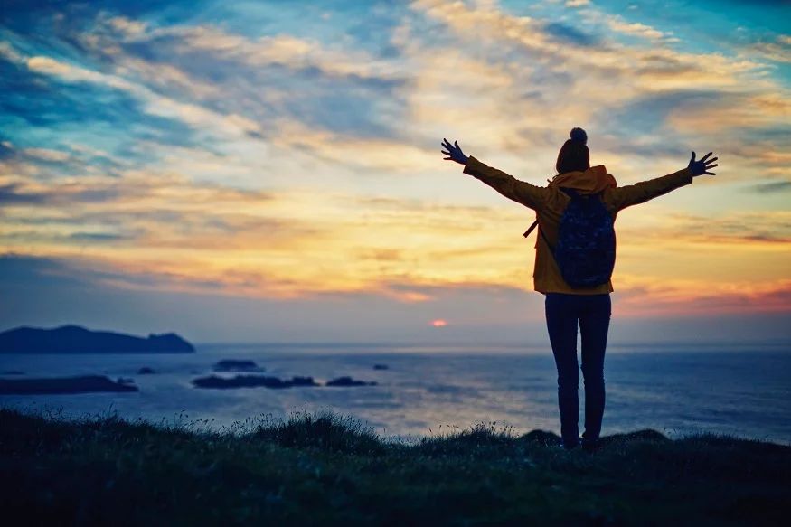 Carefree hiker standing with arms outstretched by sea during sunset